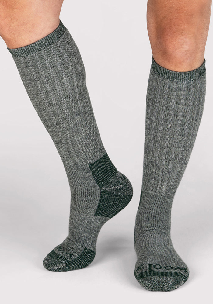 A person is wearing Woolx Mens Extreme Over The Calf Socks in Forest Dark Moss—knee-high heavyweight merino wool socks with reinforced heel, toe, and arch areas—standing on a light background.