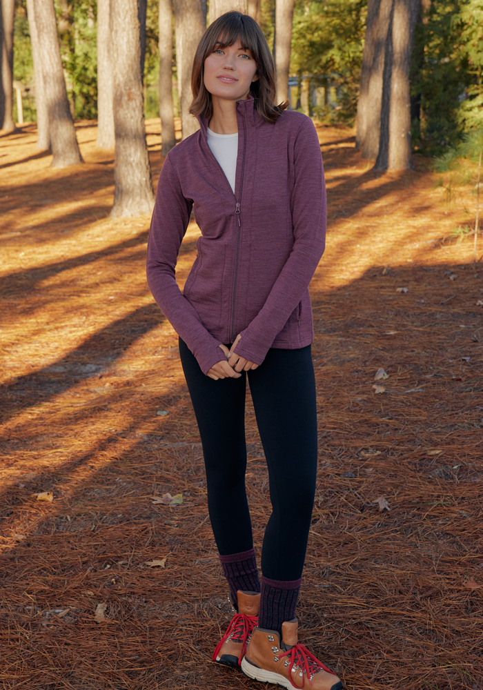 A person stands outdoors among tall pines, wearing the Woolx Finley Zip Up Sweatshirt in Grape Harvest Melange, black leggings, and brown hiking boots with red laces. Pine needles cover the ground as sunlight filters through the trees.