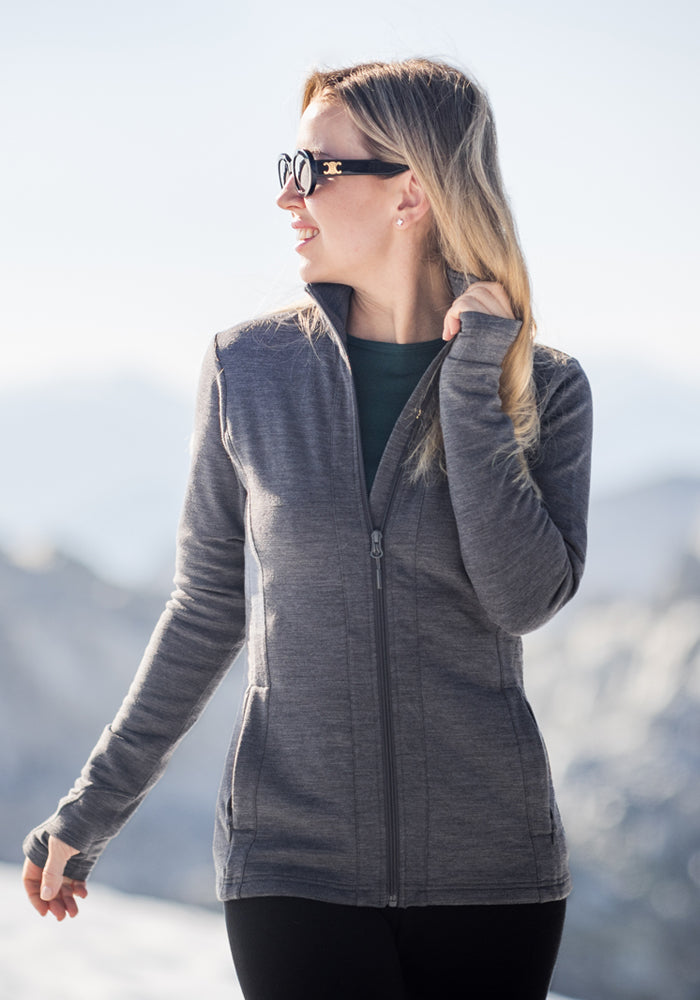 A woman in sunglasses smiles outdoors, wearing the Woolx Finley Zip Up Sweatshirt in Black. The bright, blurred background hints at a mountainous or snowy landscape.