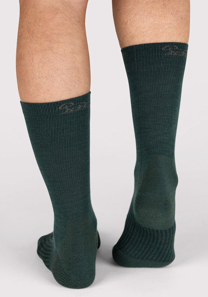 A close-up of the back of a person's lower legs wearing Woolx Mens Harbor Crew Sock in Forest, featuring moisture-wicking ribbed fabric, standing on a light-colored surface.
