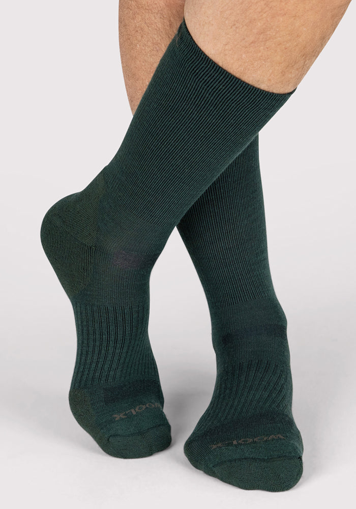 A close-up shows a person wearing Woolx Mens Harbor Crew Sock in Forest, ribbed Merino wool, feet crossed at the ankles against a plain background—ideal for moisture-wicking comfort.
