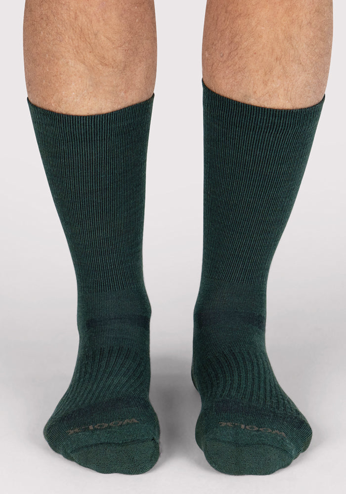A close-up of a person's lower legs wearing Woolx Mens Harbor Crew Socks in Forest, made from ribbed Merino wool with moisture-wicking properties, standing on a light surface.