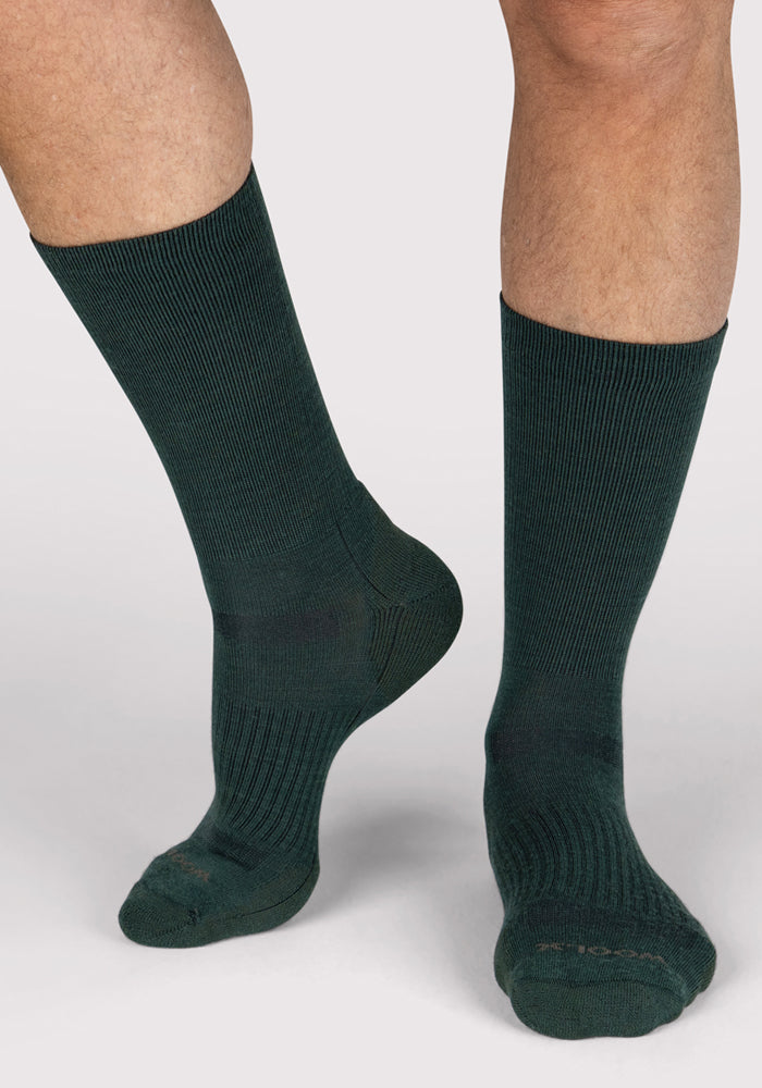 A person wears Woolx Mens Harbor Crew Socks in Forest, standing on a light surface with one foot flat and the other heel raised, showcasing the socks' temperature regulation from multiple angles.