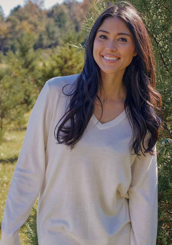 A woman with long dark hair, smiling outdoors in the sunlight with green trees behind her, wears the Woolx Francesca Sweater in Black.