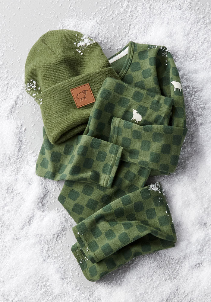 A Woolx Kids Baylor Beanie in Turtle Shell green pairs with green checkered pajamas featuring white sheep, all arranged on snow and sprinkled with delicate snowflakes.