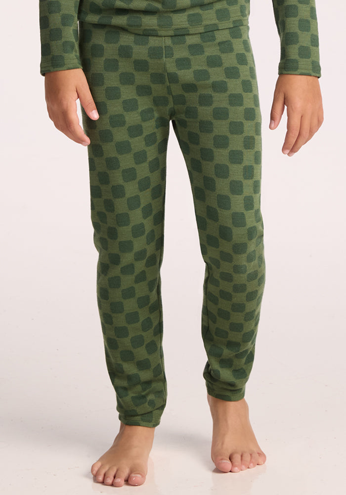 A child wearing Woolx Kids Explorer Base Layer Bottoms in Frog Hop, featuring a dark green square pattern, stands barefoot on a light surface. These cozy baselayer pants offer warmth and comfort. The upper body is partially visible. 