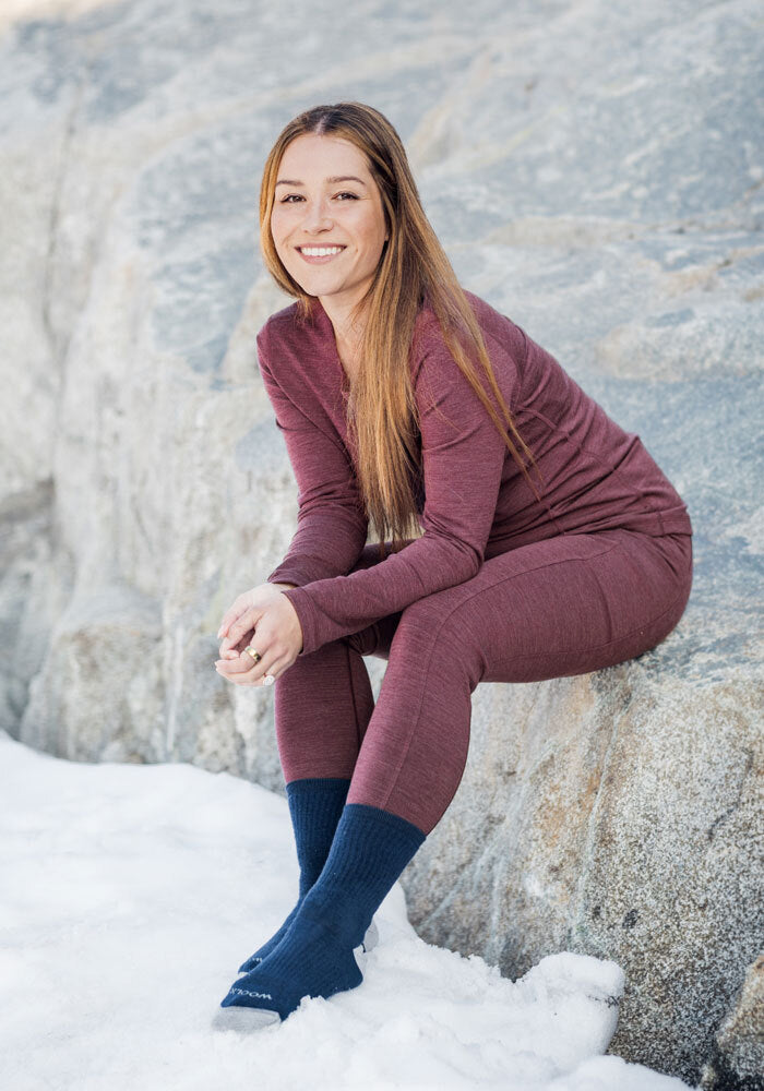 A woman with long brown hair, wearing Woolx Frost Duralite® Leggings in Grape Harvest Melange and a maroon long-sleeve top, sits on a snowy rock, smiling at the camera.