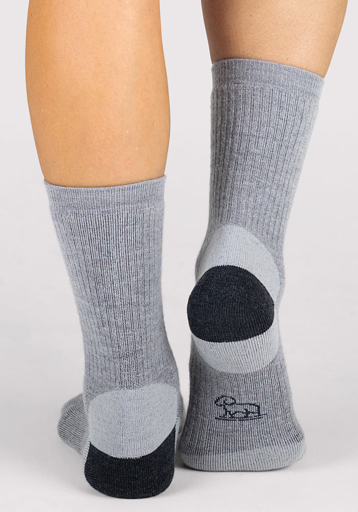 A pair of legs wears Woolx Womens Core Crew Socks - Grey. The merino wool socks are light gray with darker heels, toes, and balls, plus a small dark logo near the sole. Moisture-wicking. Plain white background.