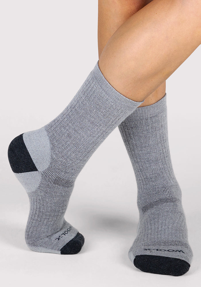 A person wearing Woolx Womens Core Crew Socks in grey, featuring odor resistance and ribbed crew style with dark gray toes and heels, stands on a light background with one foot slightly crossed in front of the other.