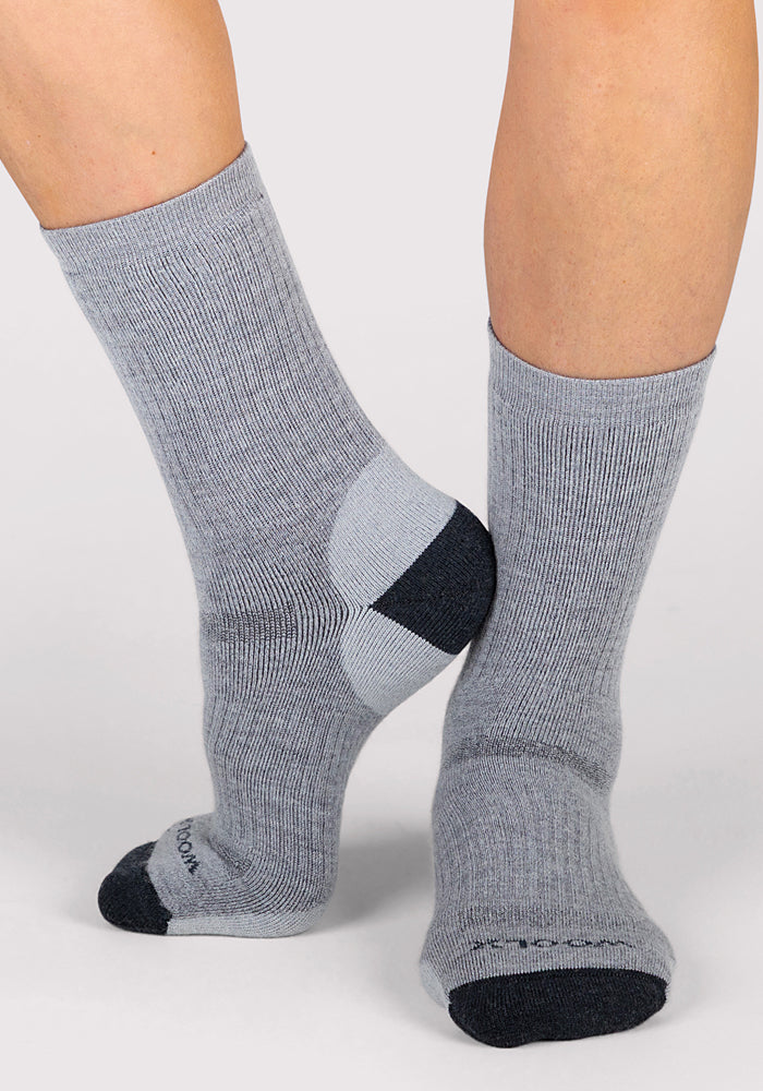 A person in Woolx Womens Core Crew Socks - Grey, featuring mid-calf merino wool with dark gray heels and toes, stands on a light background. These socks are moisture-wicking and odor-resistant for all-day comfort.