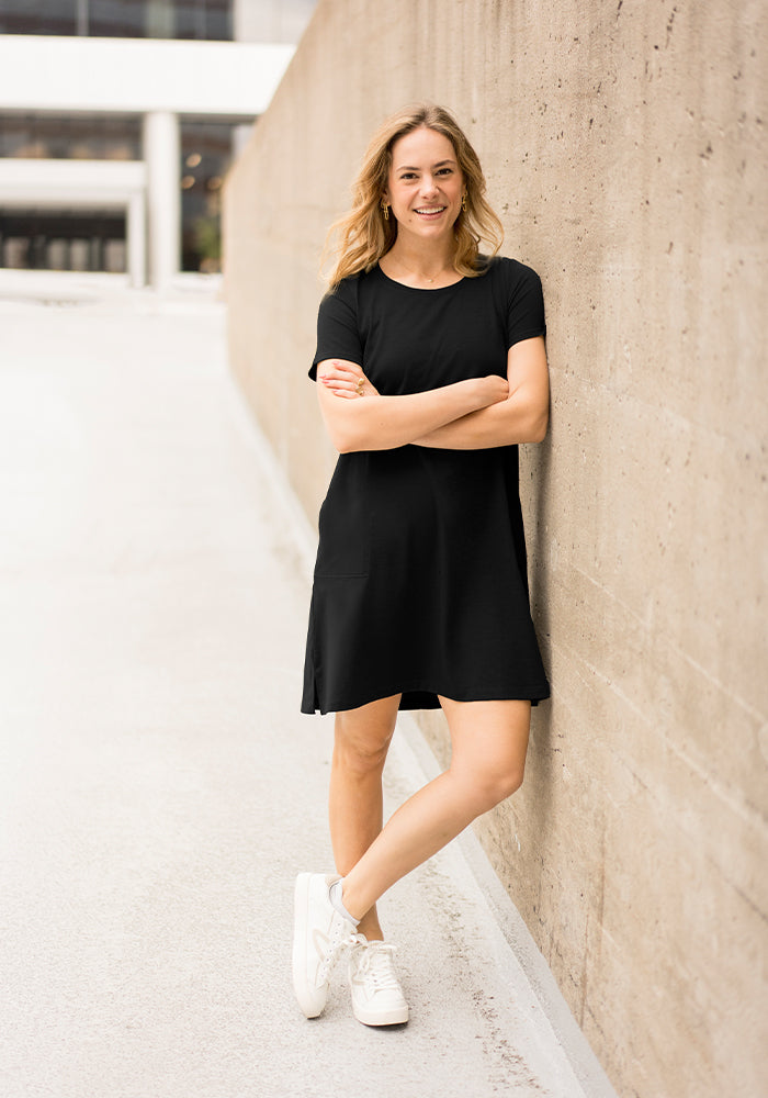Woman wearing the Woolx Georgie Dress - Black and white sneakers smiles as she leans against a concrete wall outdoors.
