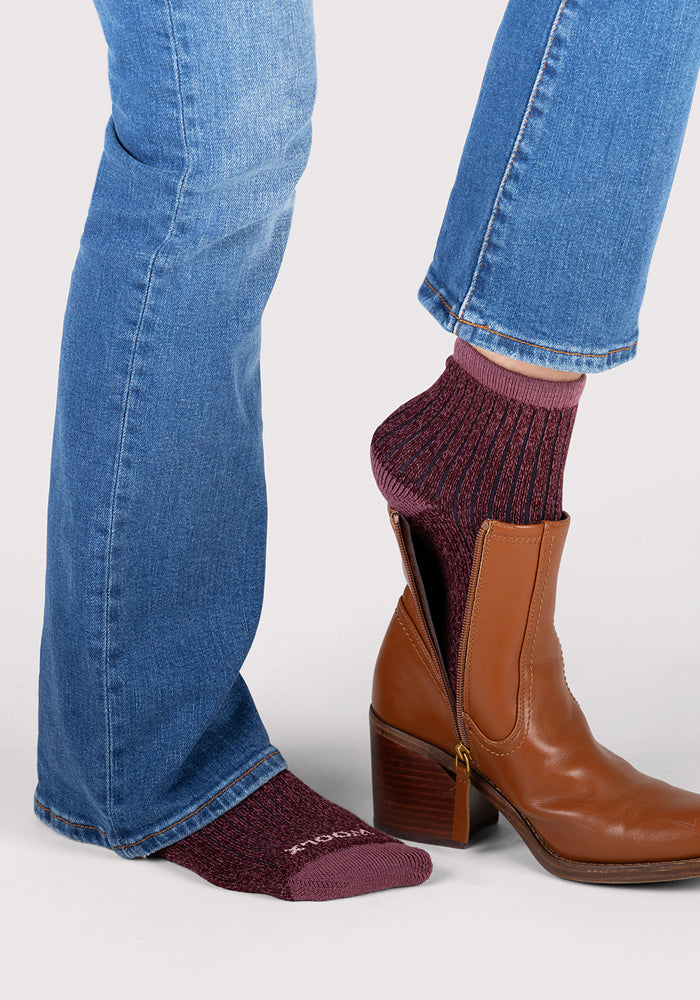 A person in blue jeans wears a brown heeled boot with a side zipper on one foot, revealing the luxury Woolx Womens Cozy Mini Crew Sock in Grape Harvest—a maroon, textured mini-crew—on the other.