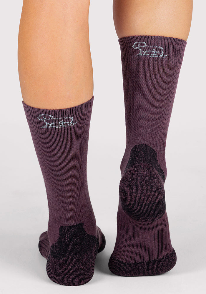 A close-up of the back of a person's lower legs wearing Woolx Womens Harbor Crew Sock in Grape Harvest, featuring moisture-wicking, breathable fabric with black heels and a white logo, standing out against a light background.