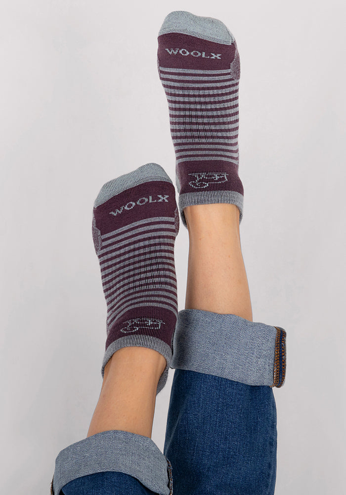 A person in rolled-up blue jeans lifts their feet, displaying Woolx Womens Amelia No Show Socks in Grape Harvest Stripe, with "WOOLX" visible on the soles. The background is plain and light-colored.