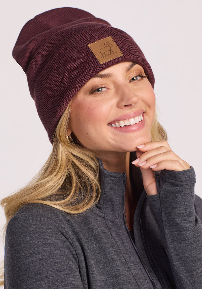 A smiling woman with long blonde hair wears the Woolx Baylor Beanie in Grape Harvest, featuring a brown patch, along with a grey zip-up jacket. She poses cheerfully with her hand near her chin against a light background.