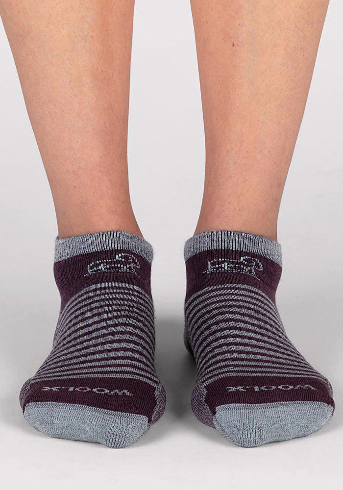 A close-up of a person’s lower legs and feet wearing Woolx Womens Amelia No Show Socks in Grape Harvest Stripe, featuring purple and gray stripes with gray toes, heels, and cuffs, standing on a light gray surface.