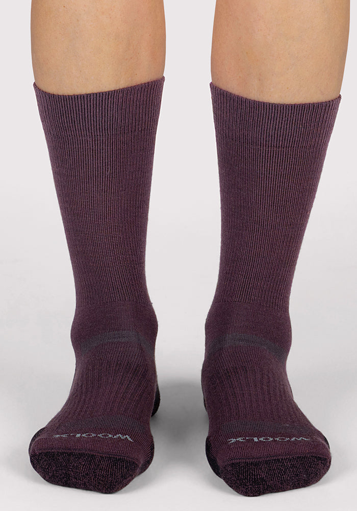 A person is wearing Woolx Womens Harbor Crew Sock in Grape Harvest, ribbed and crew-length Merino wool socks that are breathable, slightly thick, and cover the calves, standing on a light-colored surface with feet facing forward.