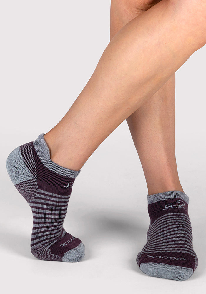 A person wearing Woolx Womens Amelia No Show Socks in Grape Harvest Stripe stands on a light gray surface with one foot slightly raised.