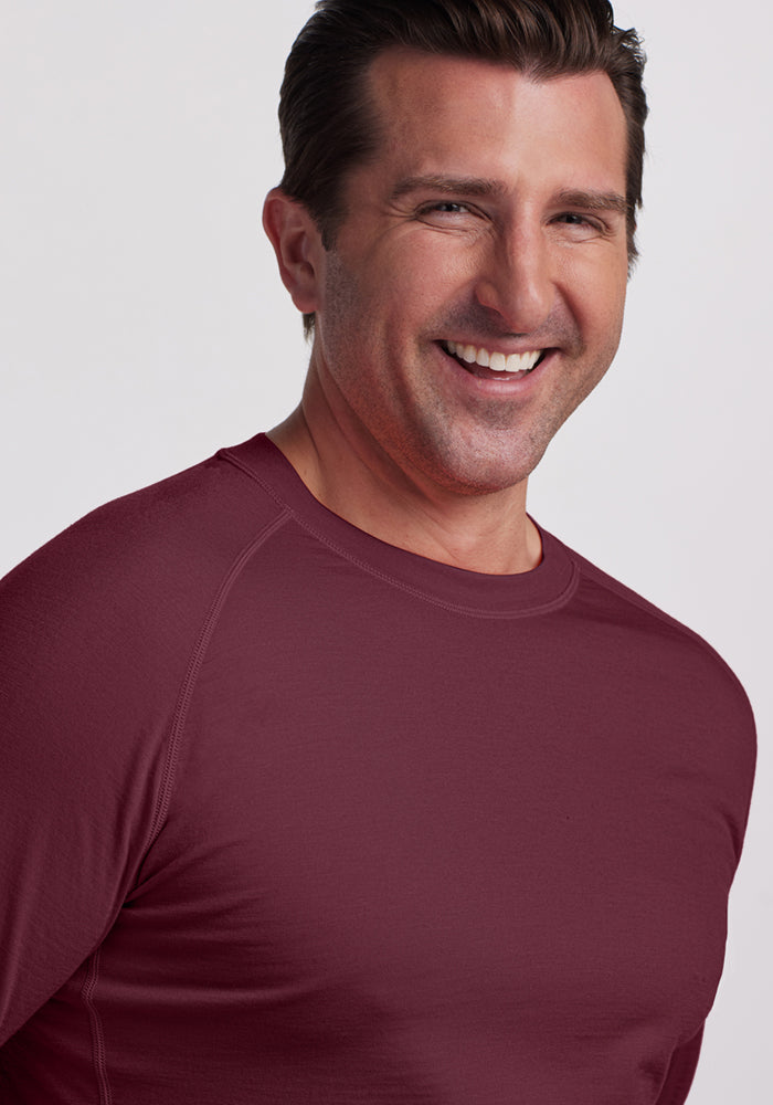 A man with short brown hair is smiling, wearing the Woolx Essential Tee - Grape Harvest, a Merino wool long sleeve known for temperature regulation, against a plain light background.