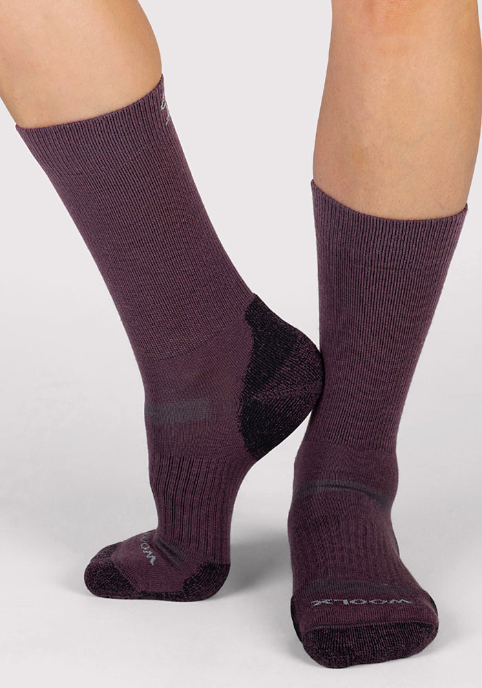 A person wearing Woolx Womens Harbor Crew Sock in Grape Harvest, featuring purple moisture-wicking fabric and black accents on the heels and toes, stands with one foot flat and the other raised on its toes.