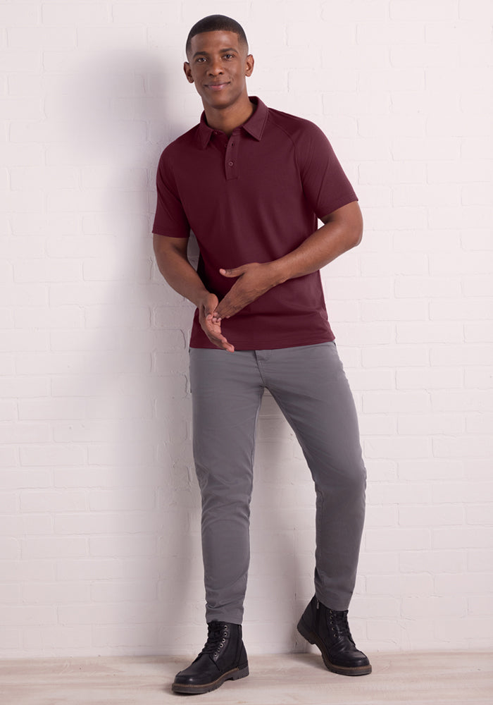A man stands against a white brick wall, smiling and gesturing while wearing the Woolx Summit Polo - Grape Harvest, paired with gray pants and black boots. He exudes a relaxed vibe and enjoys the shirt's natural moisture-wicking benefits.
