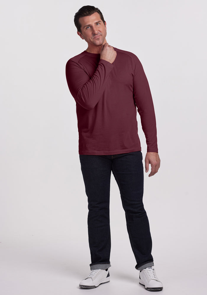 A man stands against a white background, wearing the Woolx Essential Tee in Grape Harvest, dark jeans, and white sneakers. He looks slightly upward, touching his neck, appearing thoughtful and comfortable in the temperature-regulating fabric.