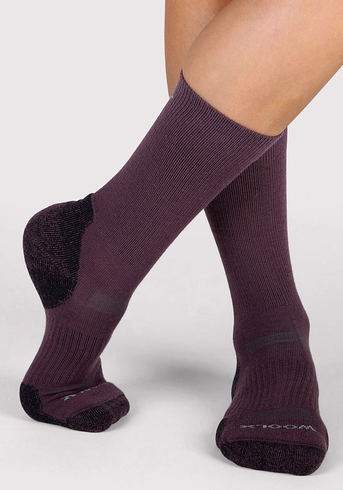 Close-up of a person’s lower legs wearing Woolx Womens Harbor Crew Sock in Grape Harvest, featuring reinforced black heels and toes. These moisture-wicking, breathable Merino wool crew socks are shown on crossed feet against a light gray background.