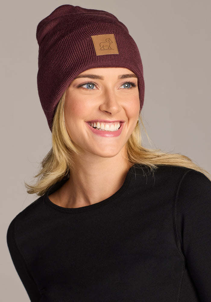 A smiling woman with blonde hair wears the Woolx Baylor Beanie - Grape Harvest, featuring a brown patch, and a black long-sleeve top, posing against a light gray background—a cozy winter hat ideal for chilly days.