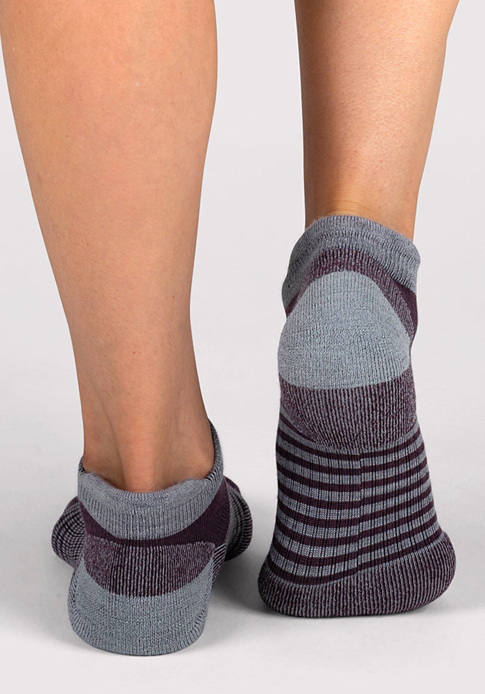 Back view of two feet in Woolx Womens Amelia No Show Socks – Grape Harvest Stripe, featuring gray and dark purple stripes with solid gray heel and toe, moisture-wicking, on a light-colored surface.