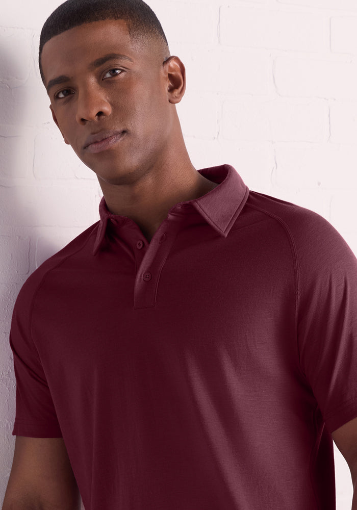 A man wearing the Woolx Summit Polo – Grape Harvest stands before a white brick wall, looking to the side with a neutral expression, highlighting the shirt’s comfort and natural moisture management.