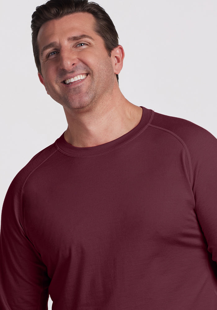 A man with short brown hair smiles while wearing the Woolx Essential Tee in Grape Harvest. The background is plain and light-colored.