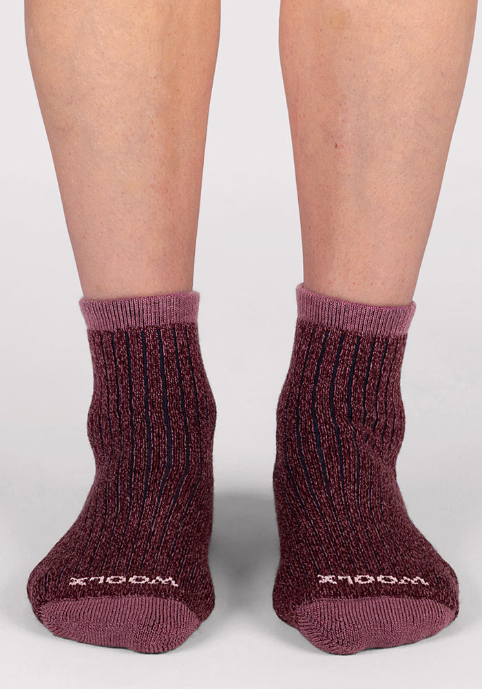 A pair of feet wears Woolx Womens Cozy Mini Crew Socks in maroon with ribbed texture, light pink cuffs and toes, and "MODEL" printed in white on the toes. The plain light gray background highlights their premium style.