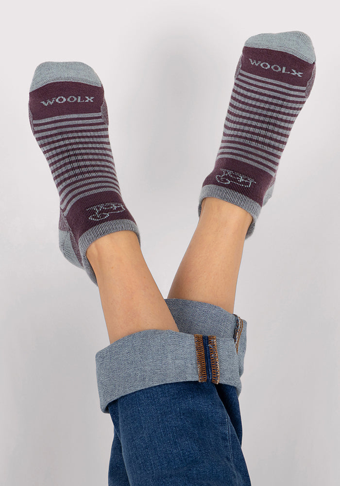 A person in rolled-up blue jeans has their feet up, revealing Woolx Womens Amelia No Show Socks in Grape Harvest Stripe. The maroon and gray striped socks feature "WOOLX" on top, set against a plain light background.
