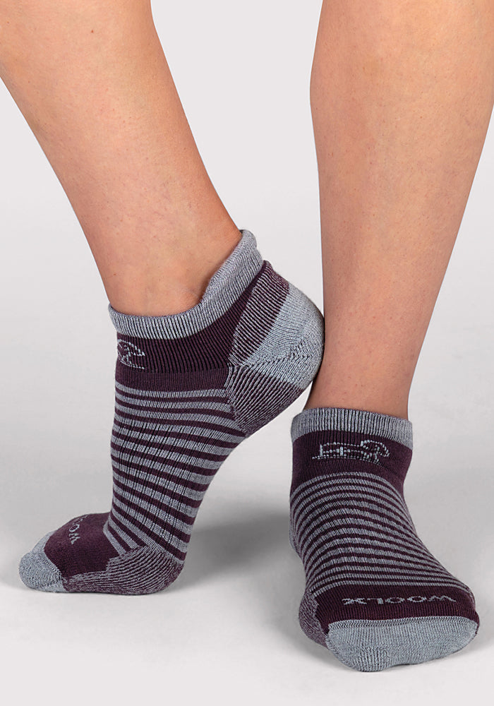 A person wears Woolx Womens Amelia No Show Socks in Grape Harvest Stripe—featuring gray and dark purple stripes with solid gray heels, toes, and cuffs—while standing on a light gray surface.