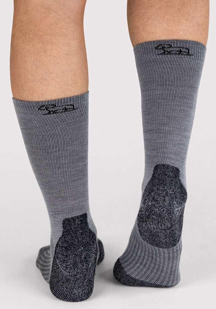 Close-up of a person's lower legs and feet wearing Woolx Mens Harbor Crew Sock - Grey, featuring ribbed Merino wool and darker heels/soles. Black text above highlights their moisture-wicking comfort.