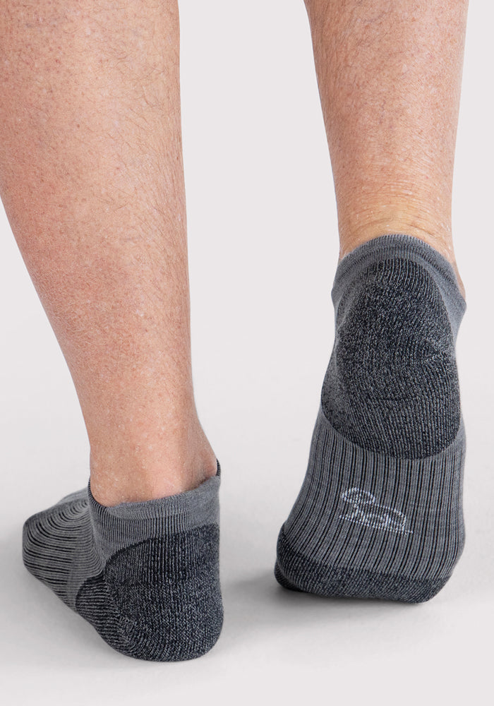 A person stands on a light background with only their lower legs and feet visible, wearing Woolx Mens Rowan No Show Socks in grey, which feature cushioned, darker toe and heel sections for added comfort.