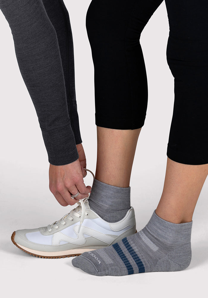 Two people are shown from the knees down; one ties a white sneaker for another, who is wearing Woolx Womens Rambler Ankle Sock - Grey with blue stripes and black leggings. The plain background is light-colored.