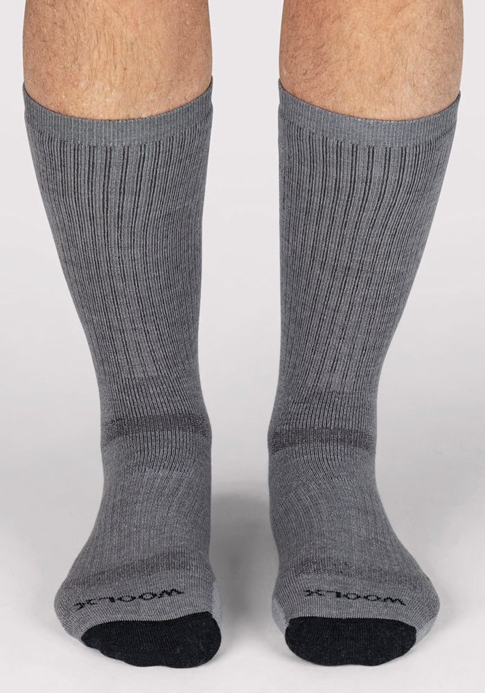 A pair of feet wearing Woolx Mens Core Crew Socks in grey, featuring moisture-wicking, odor-resistant fabric, ribbed texture, black toe areas, and small black text on the toes. Set against a plain light background.