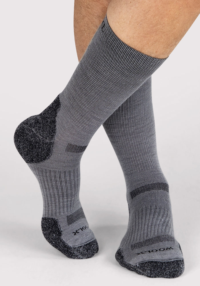 A person wears Woolx Mens Harbor Crew Socks in grey, featuring darker accents on toes, heels, and soles, standing with one foot slightly raised against a plain light background—ideal for comfort and temperature regulation.