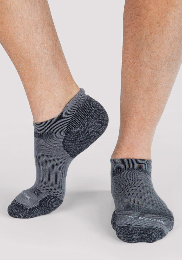 A person wearing Woolx Mens Rowan No Show Socks in Grey stands on a light background, with one foot slightly raised to showcase the side and bottom of these temperature-regulating Merino wool socks.