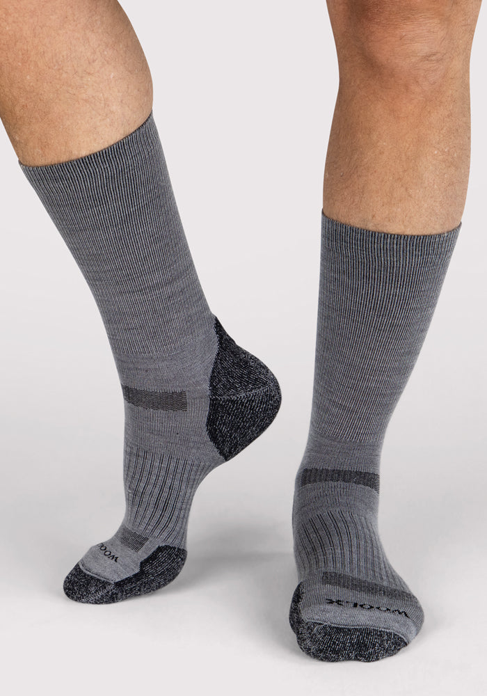 A person wears Woolx Mens Harbor Crew Socks in grey with darker reinforced heels and toes, standing on a light background. These Merino wool socks deliver temperature regulation and moisture-wicking for all-day comfort.