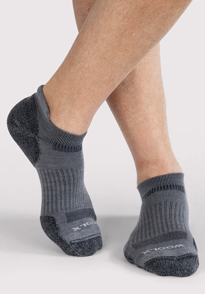 A person with crossed legs is wearing Woolx Mens Rowan No Show Socks in grey, featuring lighter ribbed sections and darker toes and heels, standing against a plain light background.