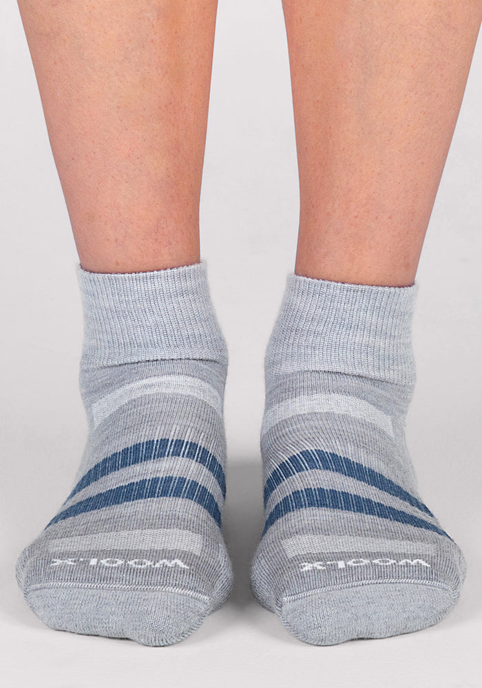 A pair of feet wearing Woolx Womens Rambler Ankle Sock - Grey, featuring moisture-wicking fabric and stylish darker gray and blue stripes, stands on a light-colored surface.