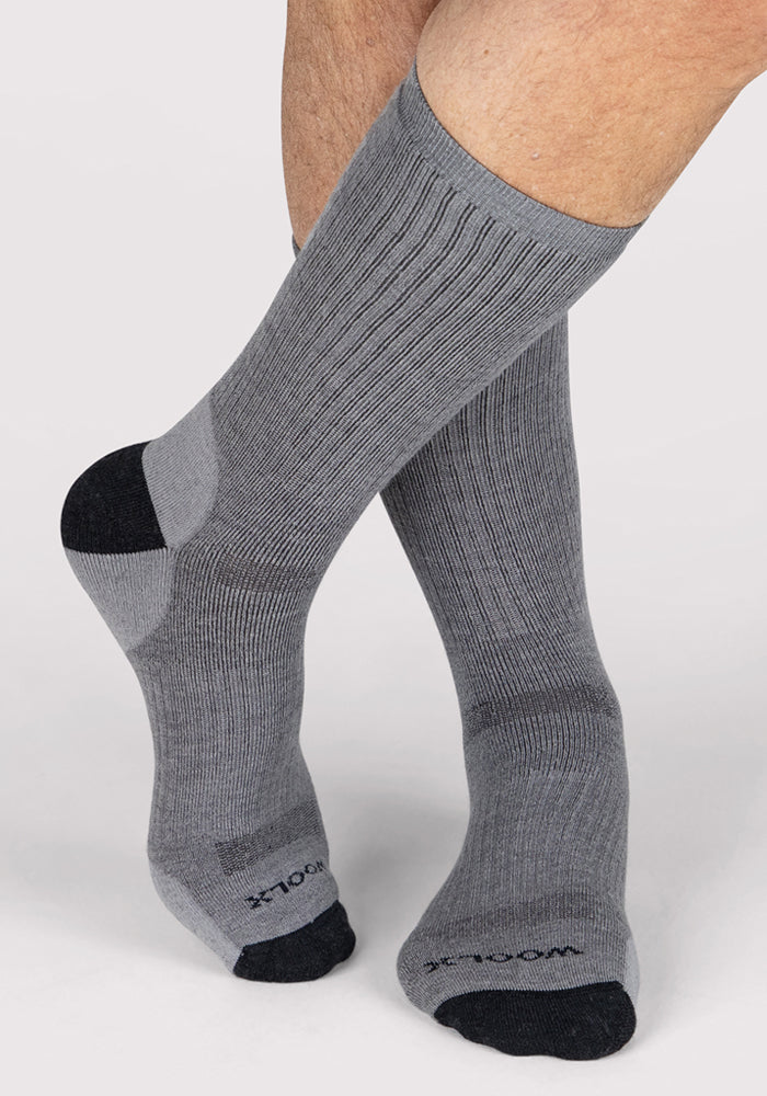 A person stands with feet crossed on a light background, wearing Woolx Mens Core Crew Socks in grey with black toes and heels. "WOOL" is visible on the toes, emphasizing the odor-resistant, moisture-wicking features.