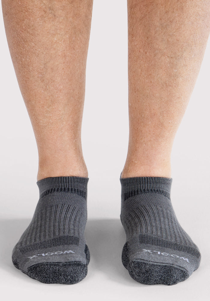 A close-up of a person wearing Woolx Mens Rowan No Show Socks in grey with ribbed detailing, shown on a light-colored surface.
