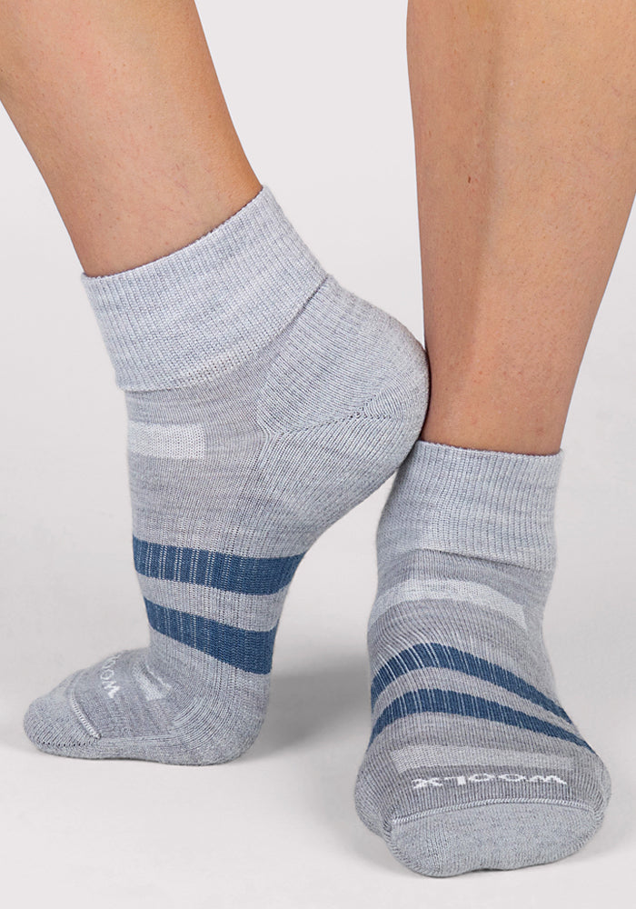A person tiptoes against a white background wearing Woolx Womens Rambler Ankle Socks in grey, featuring blue and white stripes and moisture-wicking fabric.