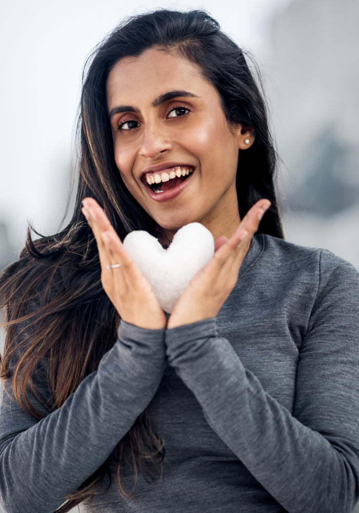 A smiling woman wearing the Woolx Sadie Baselayer Top in Deep Timber holds a heart-shaped snowball, her hair down with a blurred outdoor background.