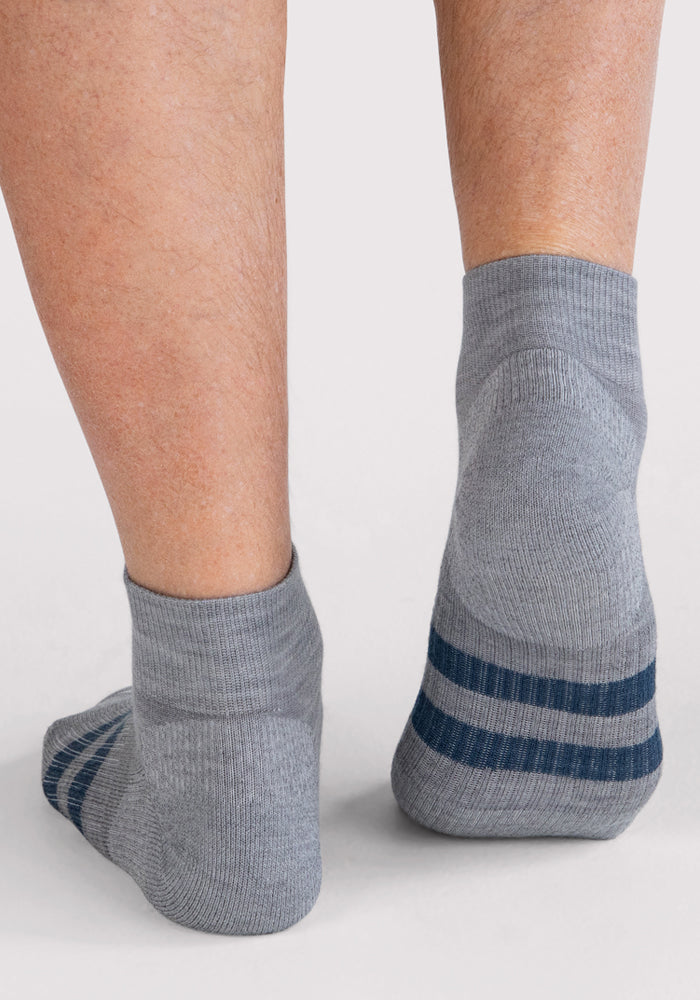 A pair of feet showcase the Woolx Mens Rambler Ankle Sock - Grey, featuring two dark blue stripes near the toes. Photographed from the back on a light background, these socks highlight their excellent moisture management properties.
