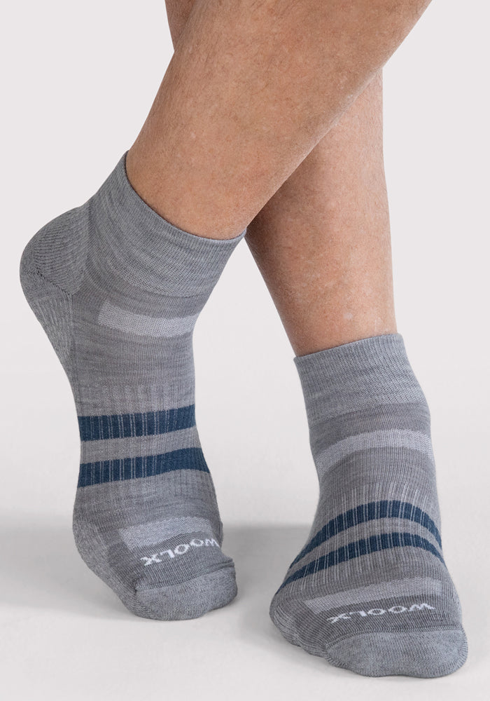 A person with crossed legs wears Woolx Mens Rambler Ankle Sock - Grey, featuring dark blue stripes and "WOOL" on the toes. These Merino wool ankle socks provide excellent moisture management, shown against a light background.