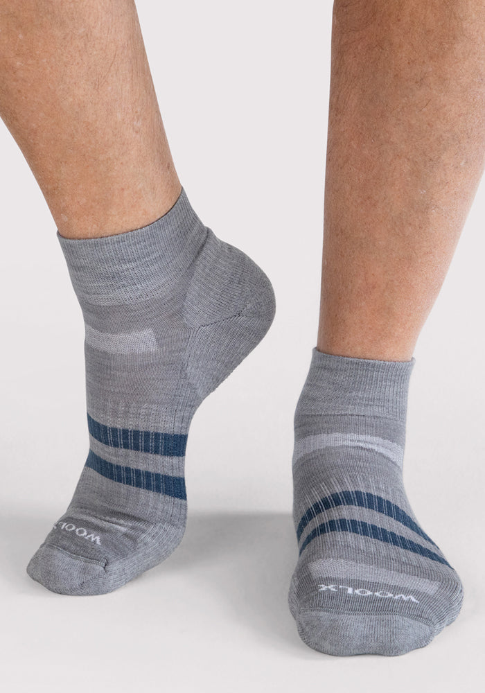 A person wears Woolx Mens Rambler Ankle Sock - Grey featuring dark blue stripes on both feet, standing on a light background. These Merino wool socks provide excellent moisture management for all-day comfort.